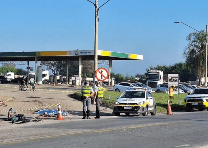 A imagem mostra a cena de um acidente fatal envolvendo um ciclista em uma rodovia. O corpo do ciclista est&aacute; coberto no asfalto, pr&oacute;ximo a uma bicicleta danificada. A &aacute;rea ao redor est&aacute; isolada com fitas zebradas amarelas e pretas, delimitada por cones laranjas. Dois policiais militares com uniformes de alta visibilidade (coletes amarelos e cal&ccedil;as escuras) est&atilde;o presentes na cena, pr&oacute;ximos a uma viatura da Pol&iacute;cia Militar. Outra caminhonete branca, da concession&aacute;ria da rodovia, tamb&eacute;m est&aacute; estacionada na via, com um funcion&aacute;rio uniformizado pr&oacute;ximo a ela. Ao fundo, observa-se um posto de gasolina e diversos ve&iacute;culos estacionados, incluindo carros e caminh&otilde;es. A rodovia de pista dupla, com tr&aacute;fego vis&iacute;vel na outra faixa. O tempo durante o dia, com boa ilumina&ccedil;&atilde;o natural. A vegeta&ccedil;&atilde;o ao redor da rodovia inclui grama e algumas &aacute;rvores. Uma placa de tr&acirc;nsito com um s&iacute;mbolo de "proibido virar &agrave; esquerda" est&aacute; vis&iacute;vel pr&oacute;xima ao posto de gasolina.