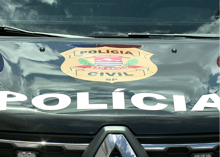 A imagem mostra a parte frontal de um ve&iacute;culo automotor, uma viatura da Pol&iacute;cia Civil do estado de S&atilde;o Paulo, no Brasil. O cap&ocirc; do ve&iacute;culo &eacute; de cor escura e possui um grande adesivo branco com a palavra "POL&Iacute;CIA" em letras mai&uacute;sculas e em negrito, centralizado na parte inferior. Acima dessa inscri&ccedil;&atilde;o, no centro do cap&ocirc;, h&aacute; um bras&atilde;o colorido. O bras&atilde;o tem um formato de escudo e cont&eacute;m diversos elementos gr&aacute;ficos e textuais. Na parte superior do bras&atilde;o, em letras pretas sobre uma faixa clara, l&ecirc;-se "POL&Iacute;CIA". Centralmente, em letras pretas sobre uma faixa clara, est&aacute; escrito "S&Atilde;O PAULO". Na parte inferior do bras&atilde;o, tamb&eacute;m em letras pretas sobre uma faixa clara, l&ecirc;-se "CIVIL" e abaixo, menor, a sigla "SP".