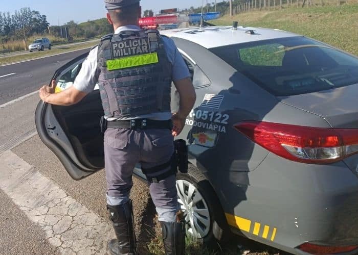 Imagem de um policial militar ao lado da uma viatura